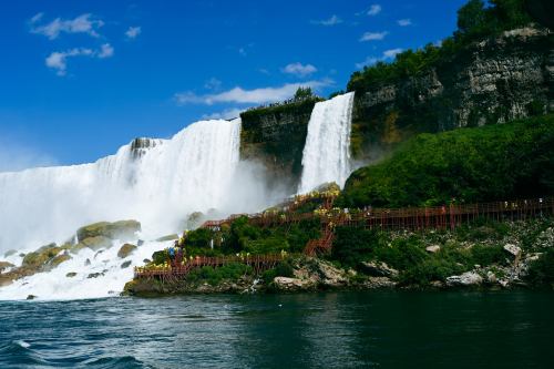 A scenic view of Niagara Falls with cascading water, lush greenery, and a wooden walkway along the riverbank.