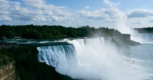 A stunning view of a waterfall cascading over cliffs, surrounded by lush greenery and a blue sky with clouds.