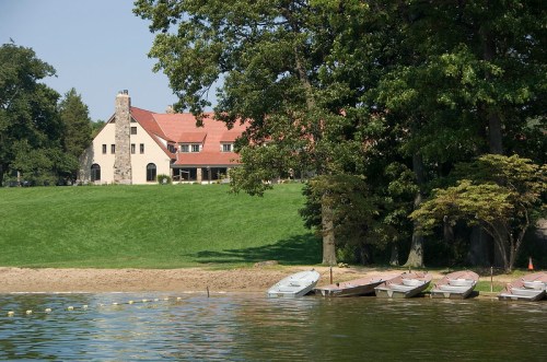 A serene lakeside view featuring a large house and several boats lined up on the shore, surrounded by lush greenery.