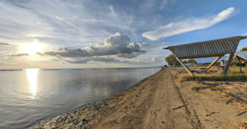 A serene beach scene at sunset, with clouds reflecting on calm water and a shelter in the foreground.