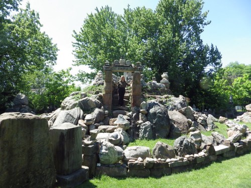 A person stands on a rocky hill surrounded by greenery, near a stone archway in a park-like setting.