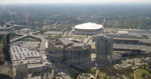 Aerial view of a cityscape featuring a stadium, parking lots, and surrounding buildings under a clear sky.