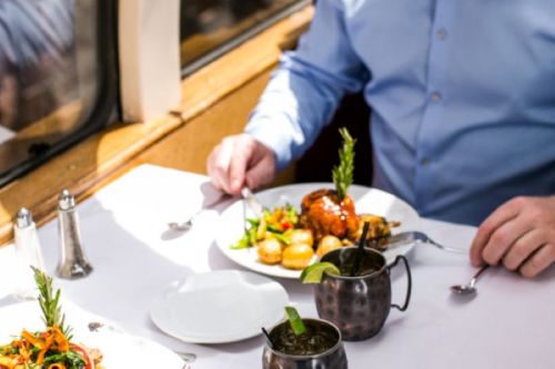 A person in a blue shirt enjoys a plated meal with vegetables and drinks on a table by a window.