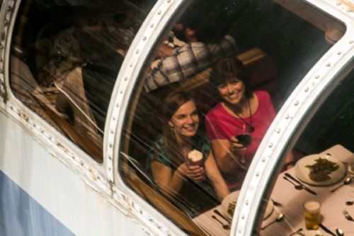 Two women smiling and holding drinks inside a restaurant with large windows, rain streaking down the glass.