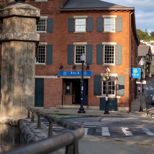 A brick building with green shutters and a blue sign reading "PALO," located on a street with lampposts and a railing.