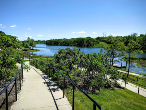 A scenic pathway leads down to a calm lake surrounded by greenery under a clear blue sky.