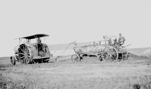 A vintage tractor pulls a plow in a field, with two men working on the equipment.