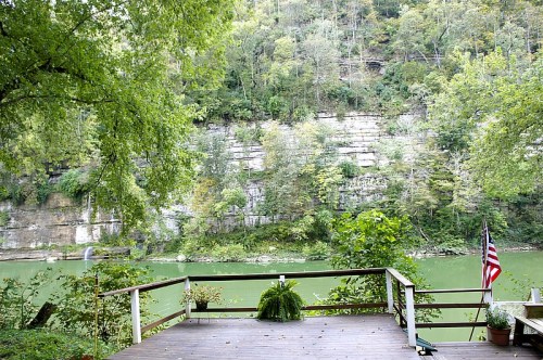 A serene riverside view with a wooden deck, lush greenery, and a cliff in the background, featuring an American flag.