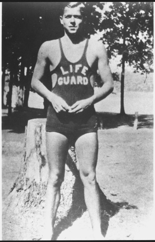 A young man in a lifeguard swimsuit stands by a tree, posing confidently outdoors.