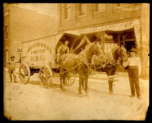 A vintage ice delivery wagon with two horses, featuring workers in period clothing, in front of a brick building.