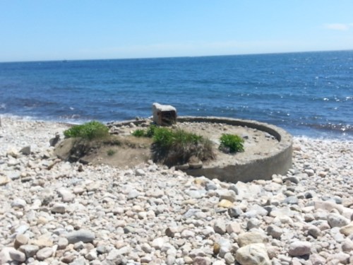 A circular stone structure surrounded by pebbles on a beach, with the ocean in the background under a clear blue sky.
