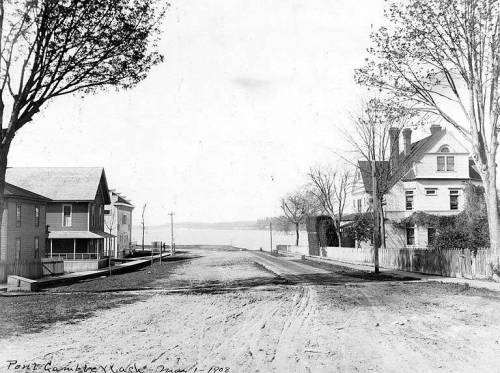 Black and white photo of a quiet street in Port Gamble, Washington, with houses and a view of the water in the background.