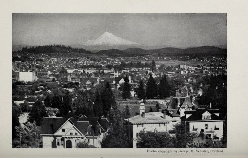 Black and white photo of a cityscape with Mount Hood in the background, featuring trees and historic buildings.