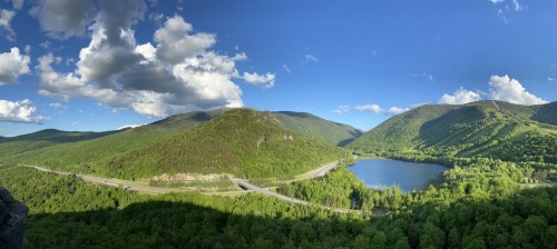 A panoramic view of lush green mountains, a winding road, and a serene lake under a blue sky with fluffy clouds.