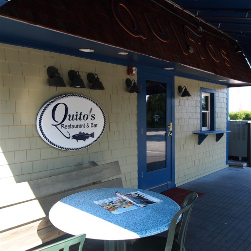 Exterior of Quito's Restaurant & Bar featuring a sign, blue door, and a small table with magazines outside.