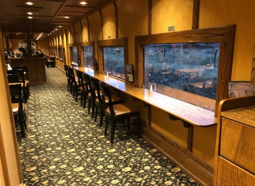 Interior of a train dining car with wooden tables, chairs, and large windows, featuring a patterned carpet.