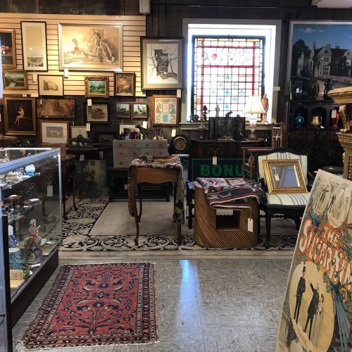 A cozy antique shop filled with framed art, vintage furniture, and decorative items, featuring a stained glass window.