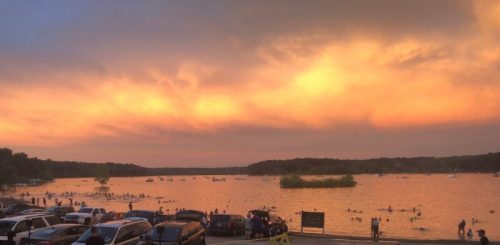 A serene lake at sunset, with boats on the water and people swimming near the shore, surrounded by trees and a colorful sky.