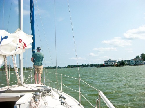 A person stands on a sailboat, looking towards a waterfront with houses and a cloudy sky in the background.
