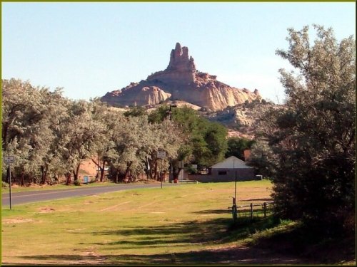 A rocky formation rises in the background, surrounded by trees and a clear blue sky, with a road in the foreground.
