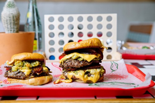 Two stacked cheeseburgers with bacon, pickles, and toppings on a red tray, with a game board in the background.