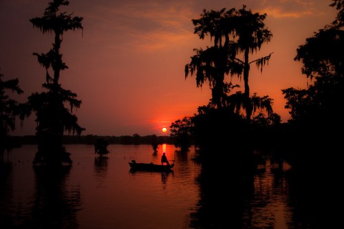 A silhouette of a person in a boat on a calm lake at sunset, surrounded by trees and a colorful sky.