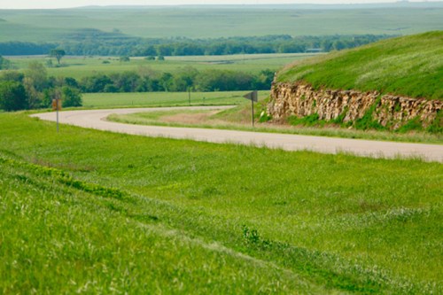 A winding road curves through lush green fields and rolling hills under a clear sky.