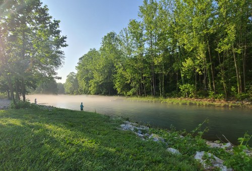 A serene river scene with mist rising, surrounded by lush green trees and a person fishing in the distance.