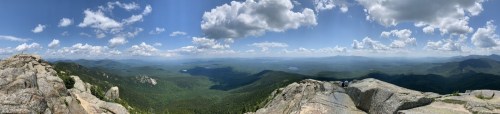 Panoramic view from a mountain peak, showcasing lush green valleys and distant mountains under a blue sky with fluffy clouds.