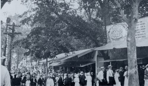 A crowded street scene from the early 20th century, with people in hats and vintage clothing near storefronts and trees.