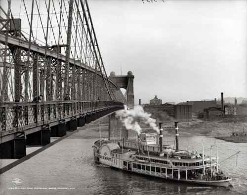 Historic black-and-white photo of a steamboat under a bridge in Cincinnati, with smoke billowing from the boat's stacks.