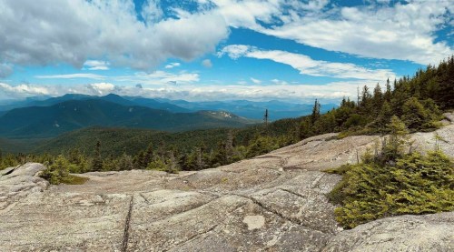 A panoramic view of mountains and valleys under a partly cloudy sky, with rocky terrain and scattered trees in the foreground.