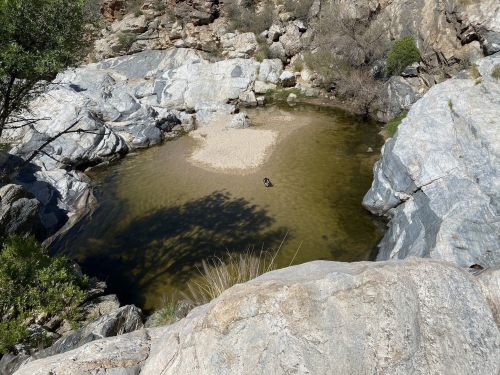 A serene natural pool surrounded by rocky terrain and sparse vegetation, with a small sandy area in the center.