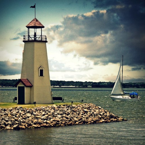 A lighthouse stands on a rocky shore with a sailboat in the background under a cloudy sky.