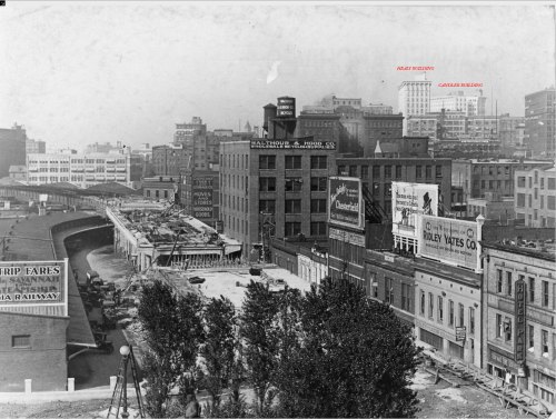 Historic black-and-white photo of a cityscape featuring buildings, a river, and advertisements along the waterfront.