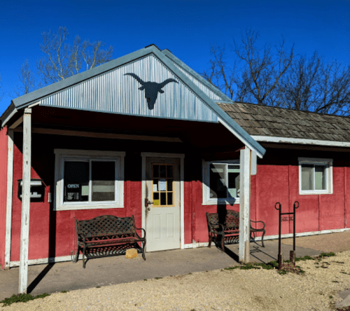 A red building with a metal roof and a longhorn silhouette, featuring benches outside and a sign that says "OPEN."