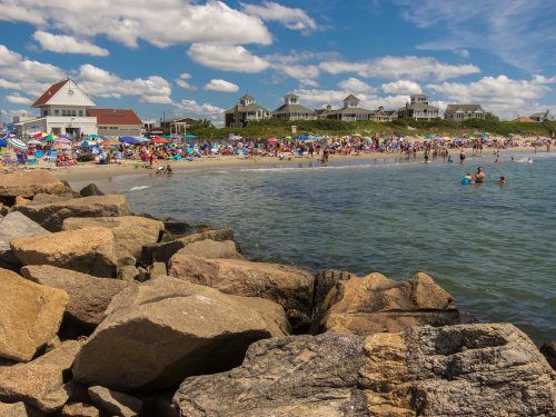 Crowded beach scene with people enjoying the water, colorful umbrellas, and rocky shoreline under a blue sky.