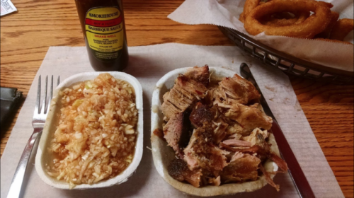 A plate of pulled pork with barbecue sauce, served with rice, alongside a bottle of barbecue sauce and onion rings.