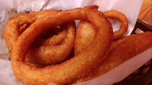 A close-up of crispy, golden-brown onion rings served in a basket lined with paper.