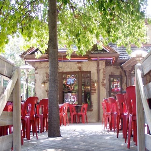 A cozy outdoor dining area with red chairs, wooden decking, and a tree, leading to a rustic building with neon signs.