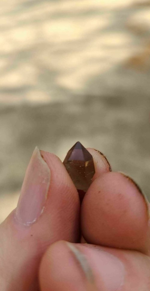A person holding a small, pointed brown crystal between their fingers against a blurred background.