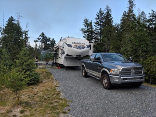 A gray truck parked next to a large RV in a forested area, surrounded by trees and gravel.