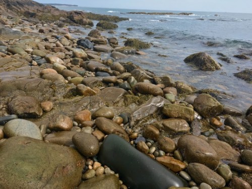 Rocky shoreline with various stones and pebbles, gentle waves lapping at the water's edge under a clear sky.