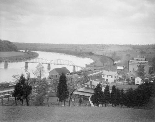 Black and white landscape featuring a river, bridge, and buildings along the shore, with trees in the foreground.