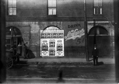 Black and white photo of a street scene with posters on a wall, a man in a suit, and a horse-drawn carriage.