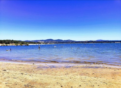 A serene beach scene with clear blue water, gentle waves, and distant mountains under a bright blue sky.