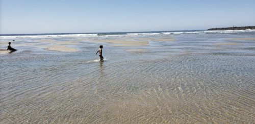 Two children play in shallow water on a sandy beach, with gentle waves and a clear blue sky in the background.
