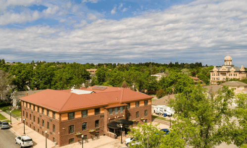 Aerial view of a brick building with a red roof, surrounded by trees and a distant clock tower under a cloudy sky.