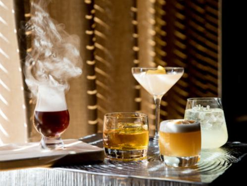 Four stylish cocktails on a table, with smoke rising from one drink, illuminated by soft light through wooden slats.