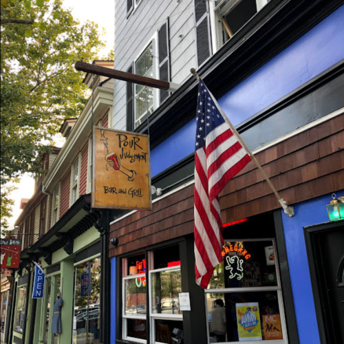 Sign for "Pour Judgment Bar and Grill" with an American flag outside a colorful building on a tree-lined street.